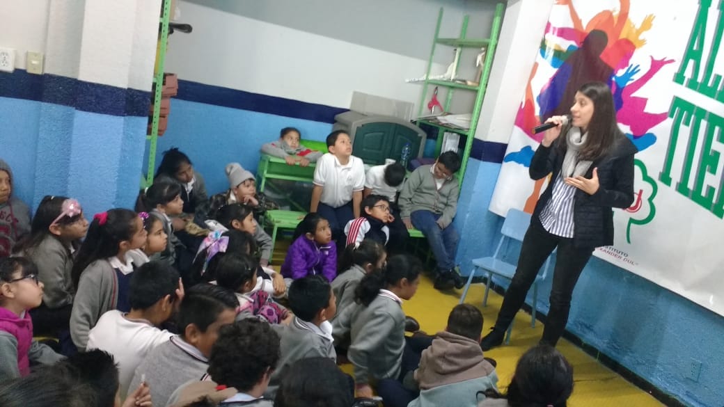 Niñas y niños sentados en el piso escuchan a una cuentacuentos en un salón del plantel Santa Lucía