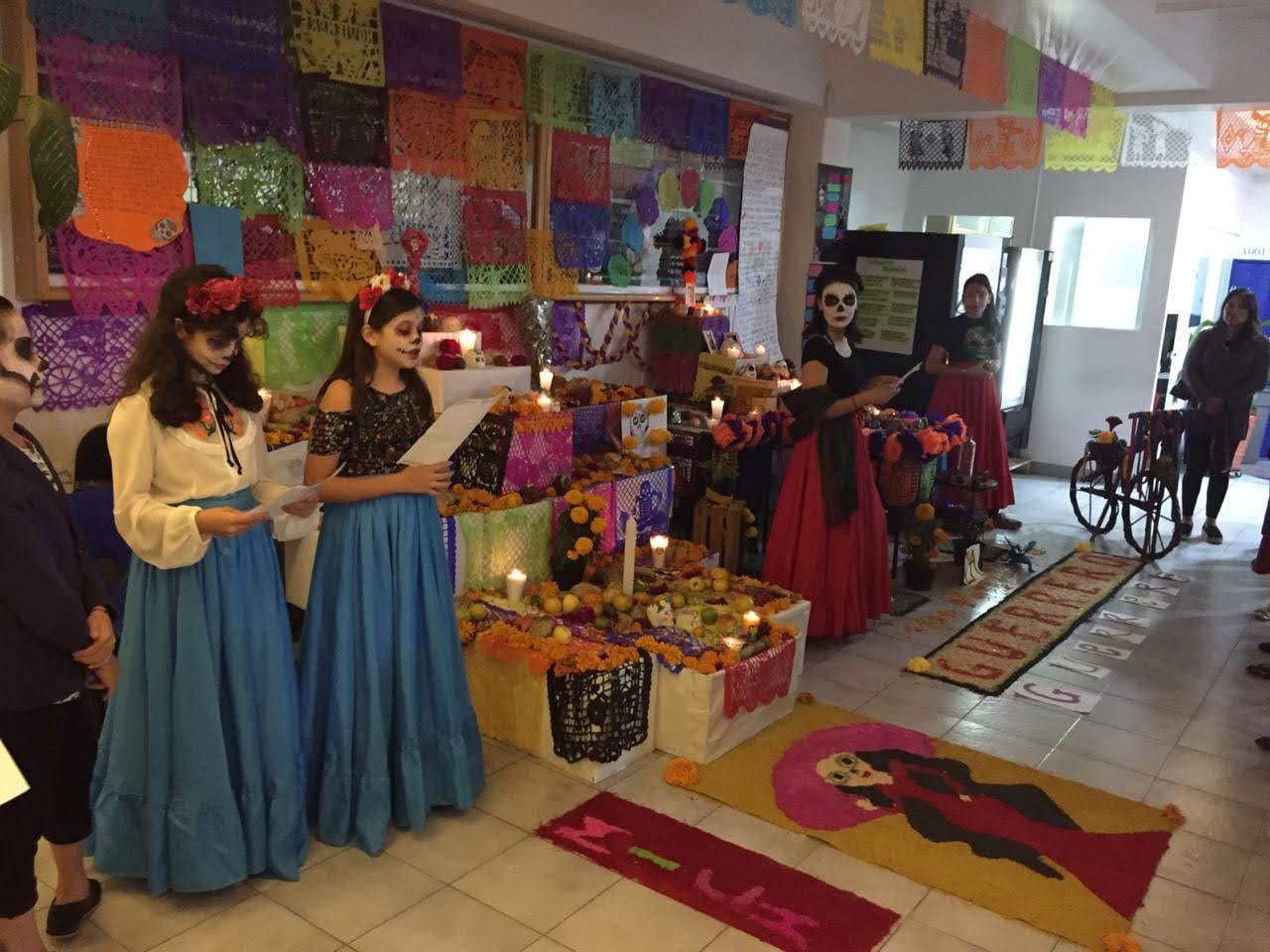 Dos alumnas con vestidos tradicionales y maquillaje de catrina leen junto a una ofrenda de Día de Muertos