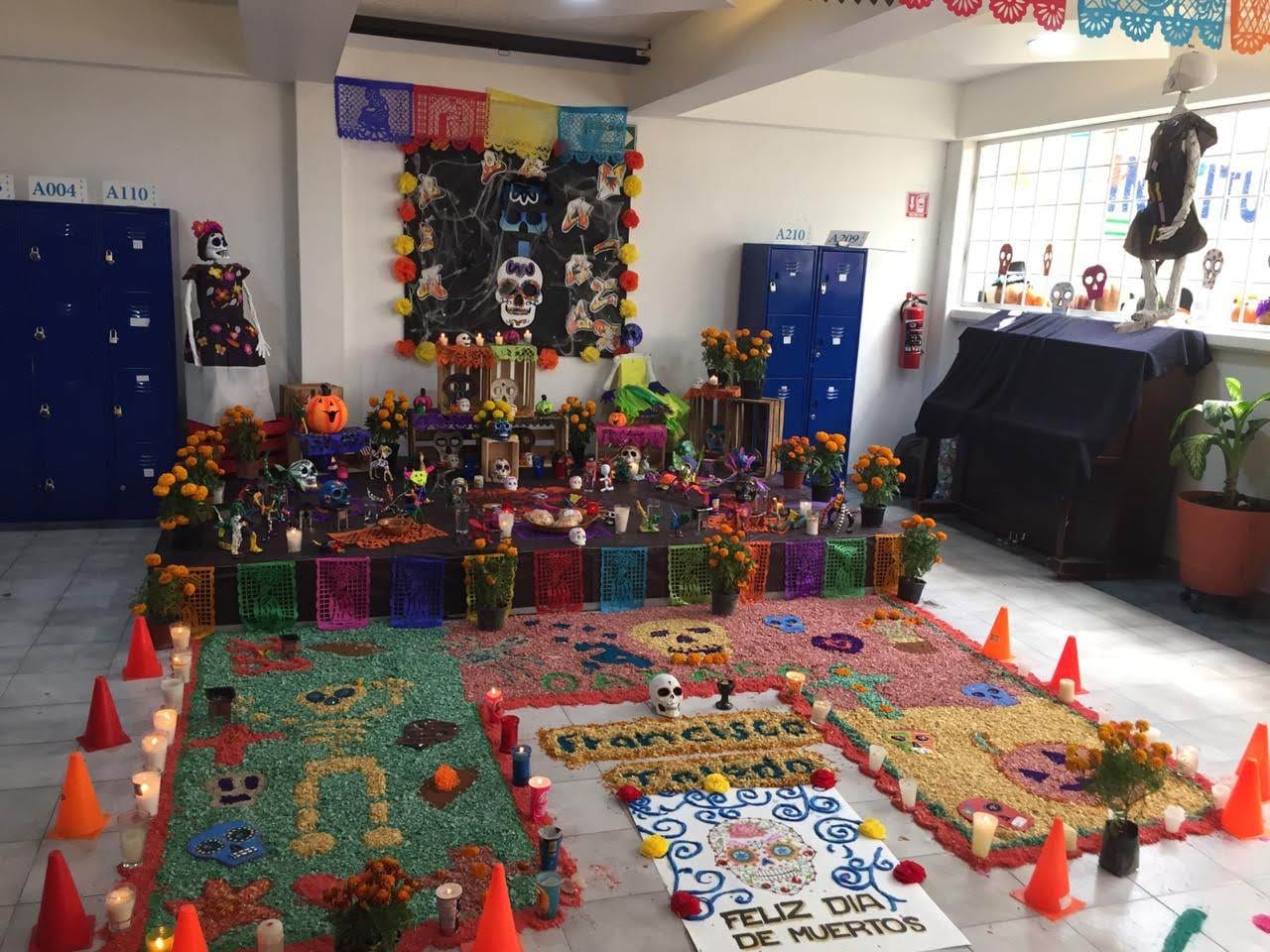 Una ofrenda de Día de Muertos con un tapete de aserrín de colores en un salón del colegio