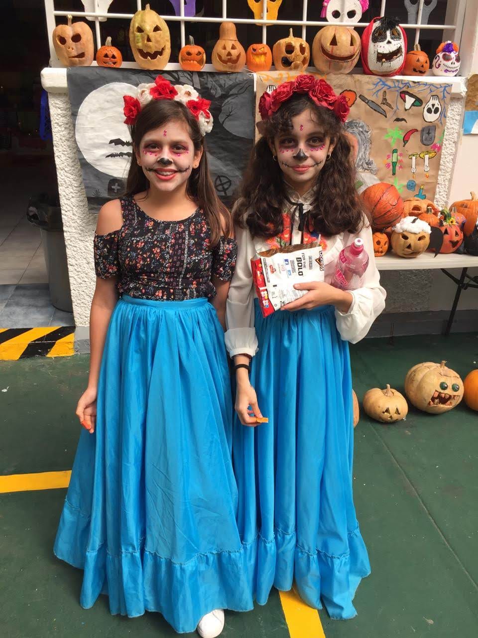 Dos alumnas con maquillaje de catrina y faldas azules posan junto a calabazas decoradas
