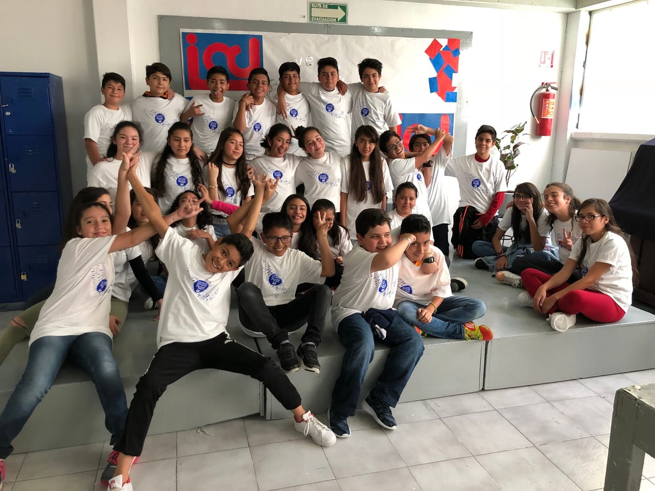 Un grupo de alumnos de secundaria con playeras blancas posa sonriente en un salón