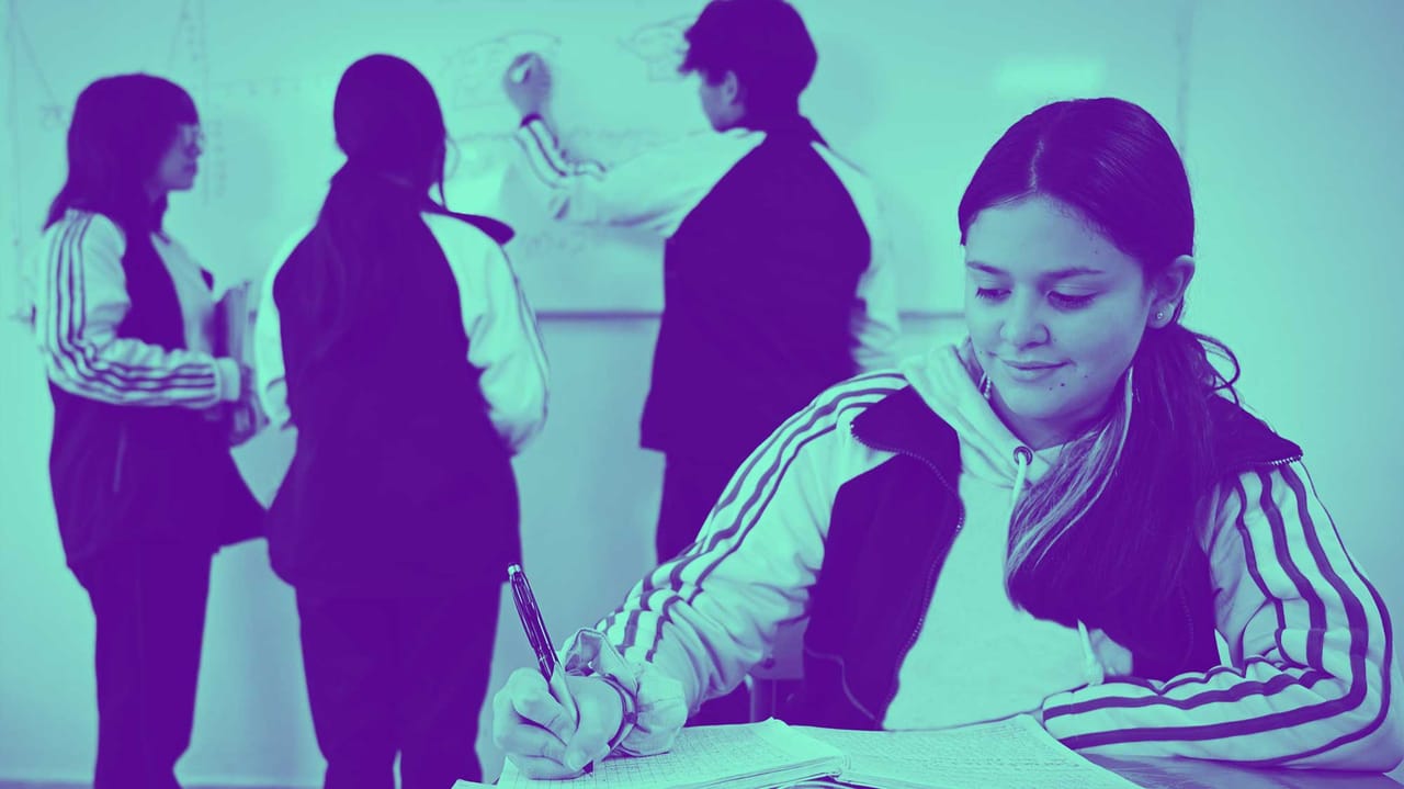 Una alumna escribe en su cuaderno mientras sus compañeros trabajan en el pizarrón